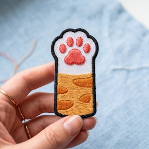 May include: Embroidered cat paw patch. The paw pad is red, with white and black borders. The lower section is orange with brown spots. The patch is held by a hand.
