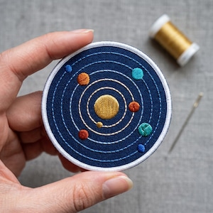 Könnte beinhalten: Gestickter Aufnäher mit einem farbenfrohen Sonnensystem-Design. Der Aufnäher hat einen dunkelblauen Hintergrund mit weißen Stichen, die die Planeten und Umlaufbahnen umreißen. Planeten in Gold-, Orange-, Blau- und Türkistönen. Der Aufnäher ist rund mit weißem Rand. Durchmesser ca. 7.6 cm.