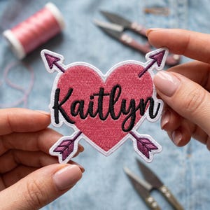 Könnte beinhalten: Ein herzförmiger Aufnäher in Rosa mit dem Namen "Kaitlyn" in schwarzer Schrift. Zwei lila Pfeile durchbohren das Herz. Der Aufnäher wird zwischen zwei Händen gehalten, im Hintergrund eine Spule rosa Garn und eine Schere.