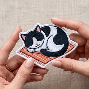 May include: Embroidered patch featuring a sleeping black and white cat on a small, orange rug. The cat has pink ears and a pink nose. The patch has a white border.