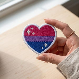 May include: Embroidered heart-shaped patch with the bisexual pride flag colours: pink, purple, and blue. The patch has a white border and includes four embroidered star accents. A symbol of LGBTQ+ pride.