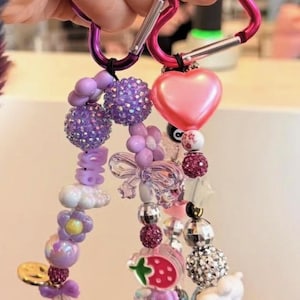 May include: A beaded charm bracelet with a pink heart-shaped charm and various beads in shades of purple, white, and silver. The bracelet is attached to a pink carabiner clip.