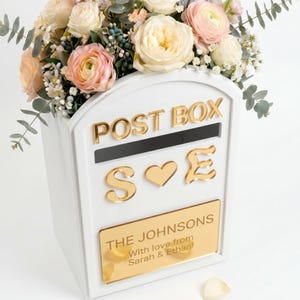 May include: A white arched post box featuring gold lettering that reads POST BOX, the letters S and E separated by a heart, and a gold plaque engraved with THE JOHNSONS, With love from Sarah & Ethan, topped with a floral arrangement of roses.