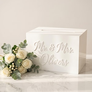 Op de afbeelding: Een rechthoekige witte acryl kaartenbox met een horizontale gleuf aan de bovenkant en de verhoogde witte tekst "Mr. & Mrs. Olivers" aan de voorzijde, vergezeld door een boeket van witte rozen, kleine witte bessen en eucalyptusbladeren.