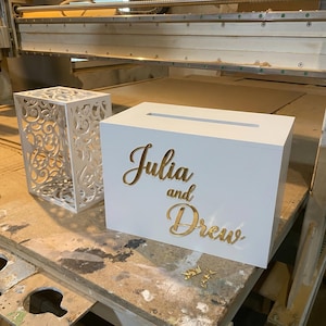 May include: A white rectangular card box with a top slot and gold script lettering reading Julia and Drew, positioned next to a white decorative box featuring an intricate laser-cut scrollwork pattern on its sides and top surface.