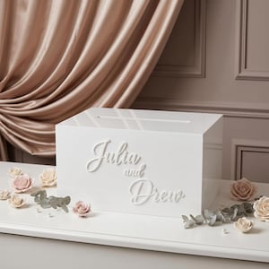 Könnte beinhalten: Eine rechteckige weiße Acrylbox mit einem horizontalen Schlitz auf der Oberseite. Die Vorderseite ist mit erhabenen weißen Schreibschriftbuchstaben versehen, die Julia and Drew lauten. Die Box ist mit kleinen künstlichen blassrosa Rosen verziert.