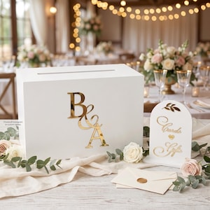 Peut inclure: Une boîte rectangulaire en bois blanc avec une fente horizontale sur le dessus et des lettres B et A en miroir doré sur le devant. À côté se trouve un panneau arqué blanc avec le texte doré Cards and Gifts et une enveloppe crème cachetée.