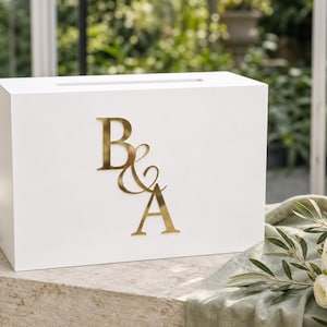 May include: A rectangular white wooden box with a horizontal slot on the top surface. The front of the box features large, metallic gold-colored letters B and A separated by an ampersand symbol, creating a personalized monogram design.