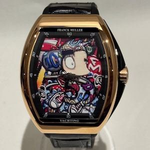 Puede incluir: Un reloj Franck Muller Yachting dorado con una correa de cuero negro. La esfera del reloj presenta un diseño de dibujos animados colorido. El reloj tiene números y agujas negros, y el texto "Franck Muller" y "Yachting" es visible.