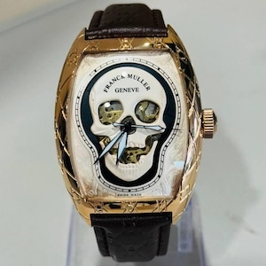 May include: A gold-tone wristwatch with a brown leather strap. The watch face features a skull design, the words "FRANCK MULLER GENEVE" and "SWISS MADE". The watch has a skeletonized design with visible internal mechanisms.