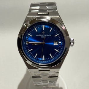 May include: A stainless steel wristwatch with a blue dial. The watch features a date display and the brand name "VACHERON CONSTANTIN GENEVE" is printed on the dial. The watch has a silver-coloured band. The watch is a luxury item.