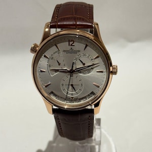 May include: A gold-toned wristwatch with a silver face and brown leather band. The watch face displays multiple subdials, hands, and the Jaeger-LeCoultre logo. The band is a dark brown, and the watch has two side buttons.