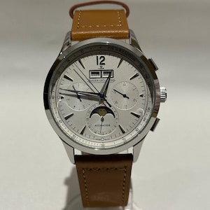 May include: A silver wristwatch with a light-coloured face, featuring subdials, a date display, and a moon phase indicator. The watch has a brown leather strap and is marked with the Jaeger-LeCoultre brand.