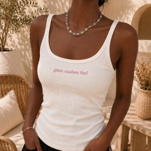 May include: A white ribbed tank top featuring a scoop neckline and thick shoulder straps. The centre of the chest displays the text your custom text embroidered in a soft pink script font. The garment is fitted and made of a textured fabric.