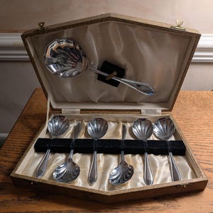 May include: A set of silver-plated spoons in a wooden presentation box. The set comprises six smaller spoons and a serving spoon, each with a shell-shaped bowl. The box is lined with satin and has a clasp to secure the contents.