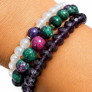 May include: A stack of three beaded bracelets. One features smooth white beads, another consists of round green and magenta speckled stone beads, and the third is made of faceted dark purple beads. The bracelets are worn together on a wrist.