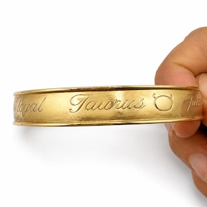 May include: A gold-toned metal bangle bracelet featuring an engraved script that reads "Loyal Taurus" followed by the Taurus zodiac symbol. The bracelet has a smooth, polished surface with raised edges along the top and bottom of the band.