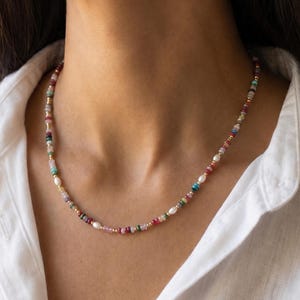 Puede incluir: Un collar de cuentas que presenta una secuencia de pequeñas rondelas de piedras preciosas multicolores intercaladas con diminutas perlas de agua dulce blancas y pequeñas cuentas espaciadoras metálicas doradas en un patrón repetitivo.