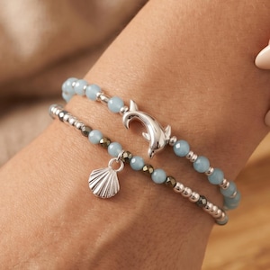 May include: Two beaded bracelets worn together. The top bracelet features light blue beads and a silver dolphin charm. The bottom bracelet consists of metallic grey beads and a silver seashell charm. Both bracelets are constructed with small round beads.