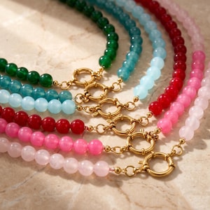 Può includere: Una collezione di cinque collane di perline disposte a ventaglio. Ogni collana presenta perline di pietra lisce e rotonde in colori distinti: verde scuro, verde acqua, azzurro, rosso e rosa. Ogni filo include una chiusura circolare color oro.