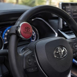 May include: A circular red textured accessory with a black Toyota logo attached to the top of a black leather steering wheel. The accessory features a metallic silver rim and is positioned on the upper left side of the steering wheel rim.