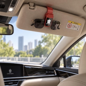 Op de afbeelding: Een rode zonnebrilhouder voor de zonneklep van een auto met het witte Crown-logo, bevestigd aan een beige zonneklep. De clip houdt een zonnebril met een zwart montuur vast. Het interieur van de auto toont een digitaal dashboarddisplay.