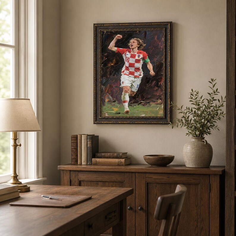 May include: An oil painting in a dark ornate frame depicts a soccer player in a red and white checkered jersey, celebrating a goal. The artwork hangs above a wooden cabinet with books and a vase.