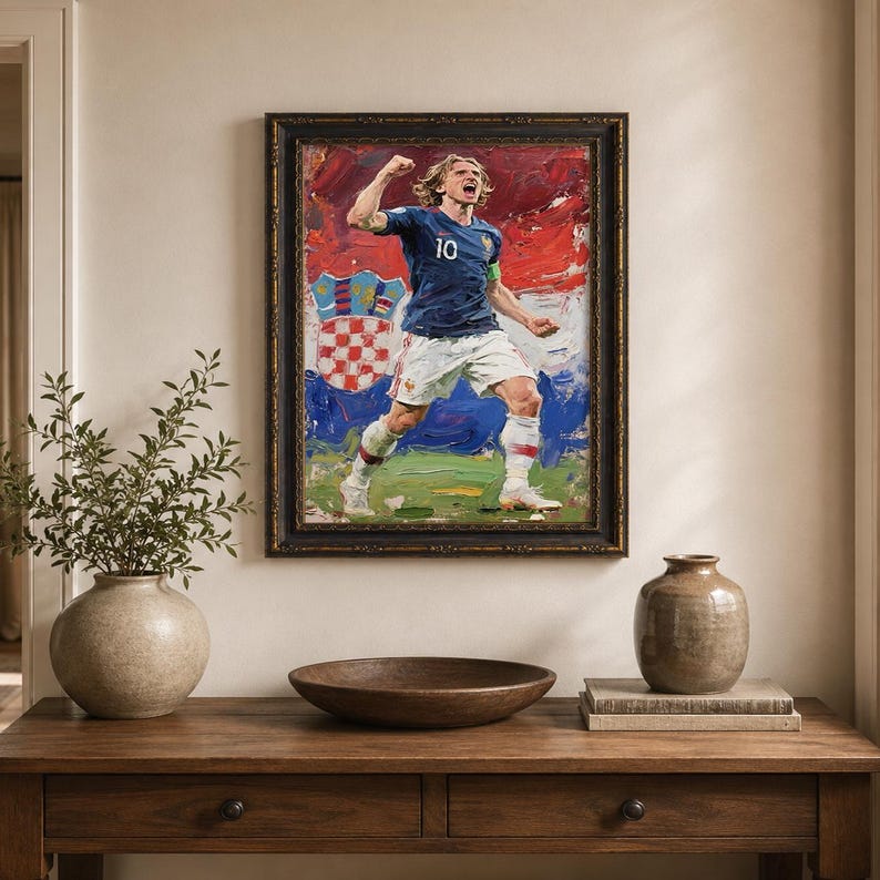 May include: Oil painting of a soccer player in a blue jersey with the number 10, celebrating a goal. The painting is set against a backdrop of red, white, and blue, framed in a dark, ornate frame. The artwork is displayed above a wooden table.