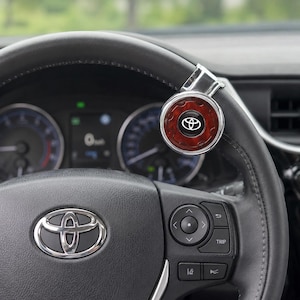 Op de afbeelding: Een ronde chromen stuurwielknop met een donkerroodbruine houtnerf-inleg en het zilveren Toyota-logo in het midden. Het accessoire is stevig bevestigd aan het bovenste gedeelte van een met zwart leer bekleed stuurwiel.