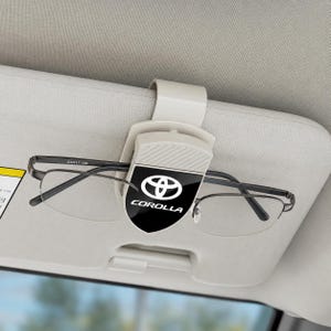 May include: A beige plastic car visor clip for spectacles featuring a black shield-shaped emblem with the white Toyota logo and the word COROLLA printed below it. The clip is attached to a light grey car sun visor holding thin-framed glasses.