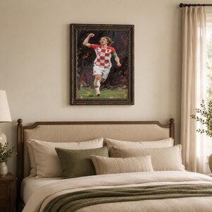 May include: Framed artwork of a soccer player in a red and white checkered jersey, celebrating a goal. The painting has a dark, textured background and a gold-toned frame. The player is running on a green field.