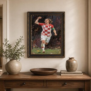 May include: An oil painting of a soccer player in a red and white checkered jersey, framed in a dark ornate frame. The player is running with his arm raised in celebration. The painting hangs above a wooden table with decorative items.