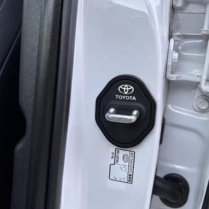 以下が含まれることがあります： 白いトヨタのロゴとテキストが施された黒いプラスチック製のドアラッチカバー。車両の白い金属製ドアフレームに取り付けられており、銀色の金属製ドアストライカーを囲んでいます。その下には、黒いテキストが印刷された小さな白い長方形のステッカーがあります。