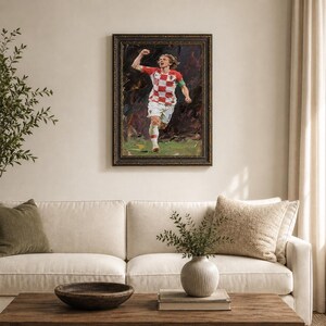 May include: An oil painting of a soccer player in a red and white checkered jersey, framed in a dark ornate frame. The player is running with his arm raised in celebration. The background is dark and abstract, with green and brown tones.