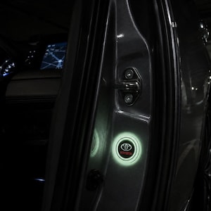 May include: A car door frame featuring an illuminated circular Toyota logo projection light. The emblem displays the black Toyota symbol and red text on a glowing white background, mounted on the interior metal panel near the door latch mechanism.