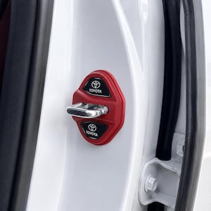 Könnte beinhalten: Eine rote Türschlossabdeckung für Autos mit zwei schwarzen Einsätzen, die das weiße Toyota-Logo und den Schriftzug tragen. Die Abdeckung ist am weißen Metalltürrahmen eines Fahrzeugs montiert und umschließt den silbernen Metalltürriegel.