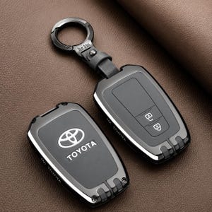 Peut inclure: Un ensemble de coques pour clé de voiture Toyota, noir et gris métallique, au design rectangulaire élégant avec un porte-clés circulaire assorti. La coque affiche le logo et le texte Toyota, avec des icônes de verrouillage et déverrouillage.