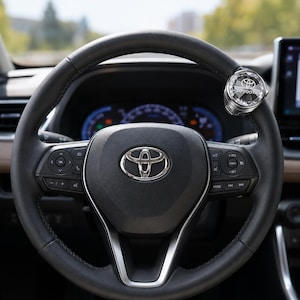 May include: A black leather-wrapped steering wheel with a central silver Toyota emblem and integrated control buttons. A circular, crystal-encrusted accessory featuring the Toyota logo is attached to the upper right rim of the steering wheel.