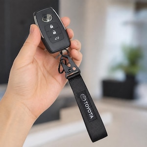 May include: A black Toyota car key fob with three buttons featuring lock, unlock, and trunk icons. The fob is attached to a black fabric strap with a white Toyota logo and text, connected by a black metal carabiner clip and a small split ring.