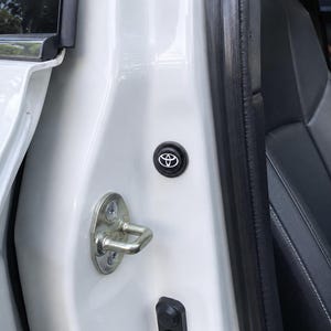 May include: A white car door frame interior featuring a circular black emblem with a white Toyota logo. Below the emblem is a metallic silver door latch mechanism secured with two screws, set against the white painted metal of the vehicle body.