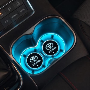 May include: Two circular black cup holder inserts featuring the white Toyota logo and text, illuminated by a bright blue LED light ring. These automotive accessories are positioned within the centre console of a vehicle near drive mode buttons.