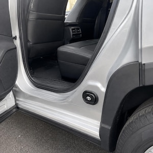 May include: The open rear passenger door of a silver vehicle reveals a black leather interior with a black floor mat. A small, circular black emblem featuring the white Toyota logo is mounted on the lower silver door frame near the rear wheel well.