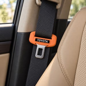 May include: A black seat belt strap featuring an orange protective cover with the word TOYOTA printed in white letters. The cover is positioned around the metal seat belt buckle tongue, which is attached to the vertical black fabric webbing.