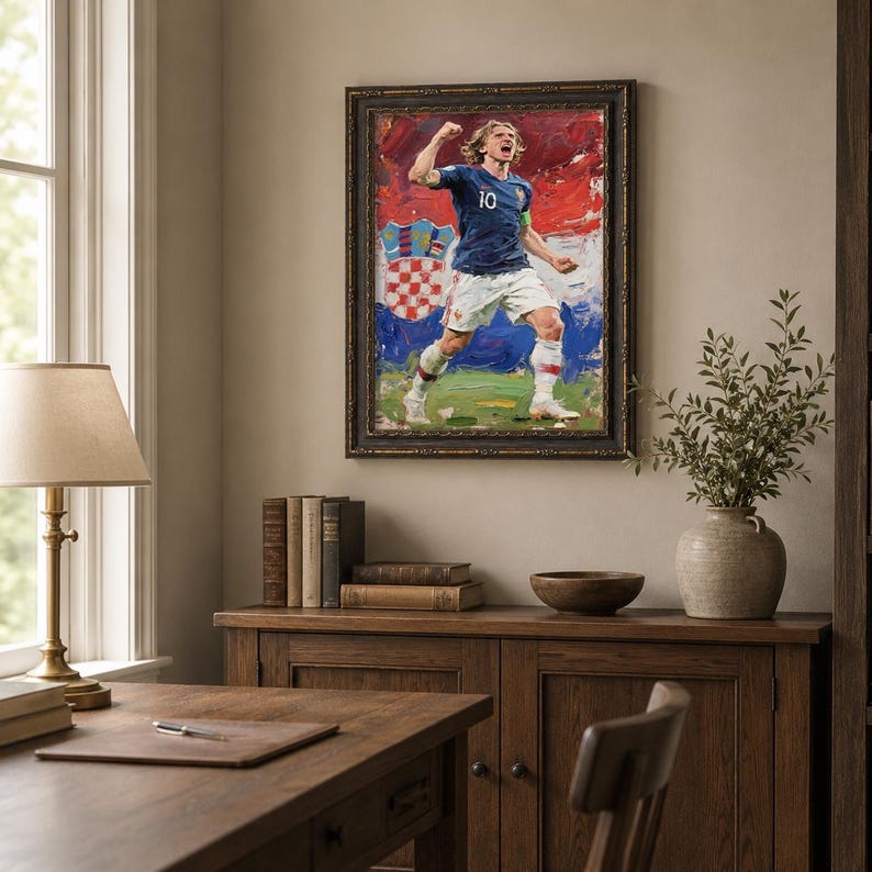 May include: A framed painting of a soccer player in a blue jersey with the number 10, celebrating a goal. The background features the Croatian flag colors. The artwork is displayed in a room with wooden furniture.