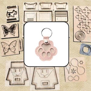 Puede incluir: Un llavero de cuero rosa claro con forma de huella con una anilla de metal plateado y un botón de presión plateado central. El llavero está rodeado por varios troqueles de madera con formas como mariposas, flores y patrones geométricos.