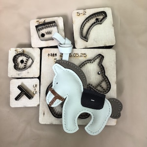 Puede incluir: Un colgante de caballo de cuero azul claro con brida marrón y silla negra, con un lazo de cuero. Se muestra sobre varios troqueles de madera etiquetados 5-2, 5-3, 5-4 y 5-5, con uno de ellos mostrando el texto 小马挂件 2016.05.25.