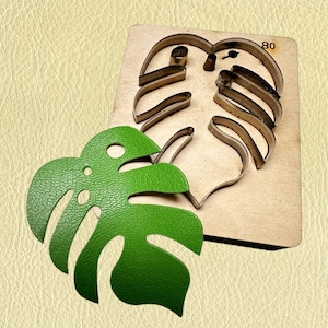 Puede incluir: Un troquel de metal con forma de hoja de monstera montado sobre una base de madera cuadrada con el número 80 impreso, acompañado de un recorte de piel sintética verde texturizada con forma de hoja de monstera con varios orificios circulares.