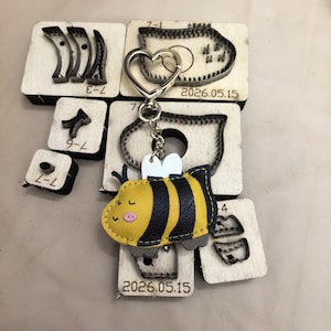 May include: A yellow and black faux leather bee keychain with white wings and a heart-shaped metal ring, resting on several wooden cutting dies labeled with numbers and the date 2026.05.15. The bee has stitched details and a pink cheek.