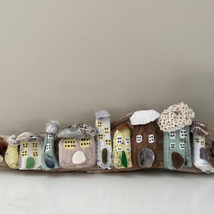 Puede incluir: Una caprichosa obra de arte en madera flotante que representa un pueblo en miniatura de casas pintadas y estructuras con forma de hongo. Las casas están adornadas con detalles coloridos, incluyendo acentos verdes, amarillos y azules, dispuestos a lo largo de un trozo de madera flotante.