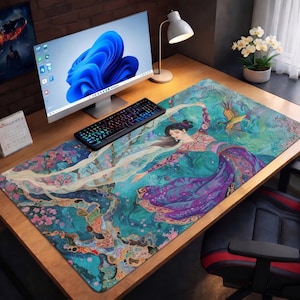 May include: A large desk mat featuring a vibrant illustration of a woman in a flowing gown, with a bird, set against a teal and purple background. A computer keyboard and monitor sit on the wooden desk, along with a desk lamp and a calendar.